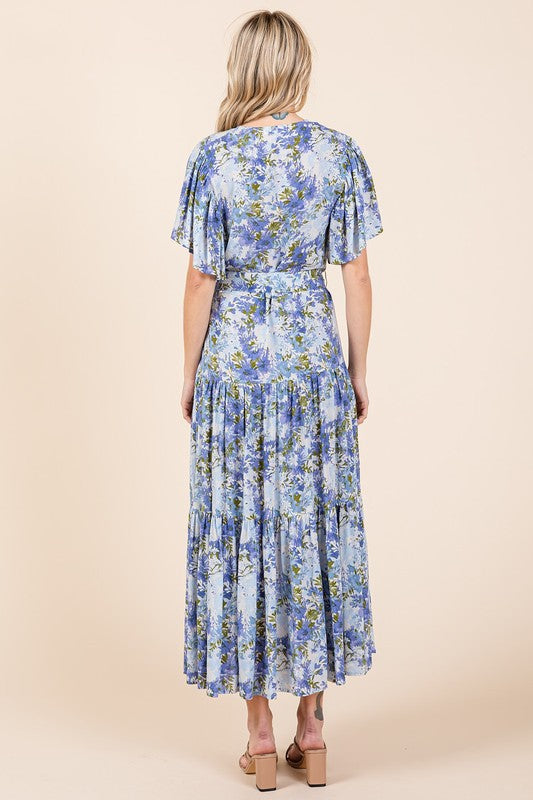 Back view of a woman wearing a floral tie-waist tiered dress with flutter sleeves, styled with neutral sandals, showcasing a flowy and feminine silhouette.