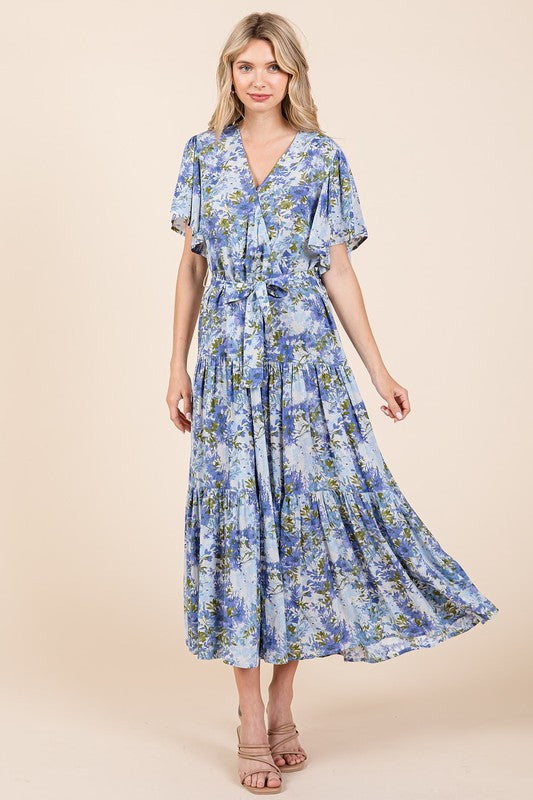 A woman wearing a floral tie-waist tiered dress with flutter sleeves, styled with neutral sandals, showcasing a flowy and feminine silhouette.