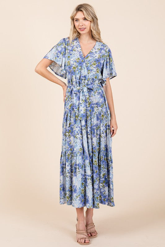 A woman wearing a floral tie-waist tiered dress with flutter sleeves, styled with neutral sandals, showcasing a flowy and feminine silhouette.