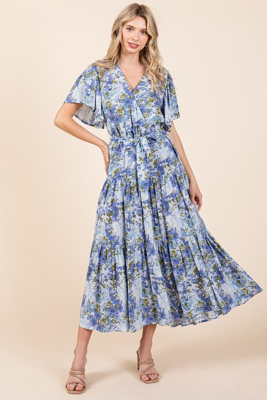 A woman with her hand on her hip wearing a floral tie-waist tiered dress with flutter sleeves, styled with neutral sandals, showcasing a flowy and feminine silhouette.