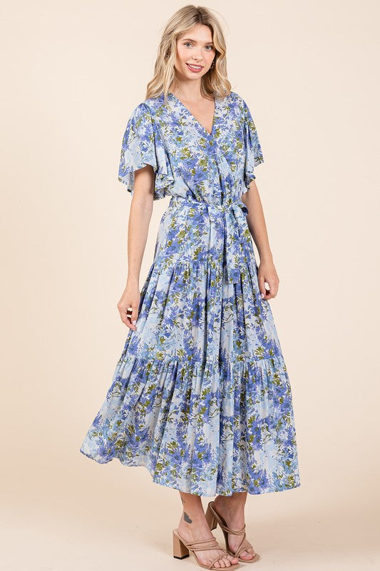 A woman wearing a floral tie-waist tiered dress with flutter sleeves, styled with neutral sandals, showcasing a flowy and feminine silhouette.