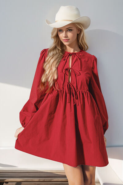 A woman wearing a red tie-front mini dress with long puff sleeves, styled with a white cowboy hat, standing in natural light.