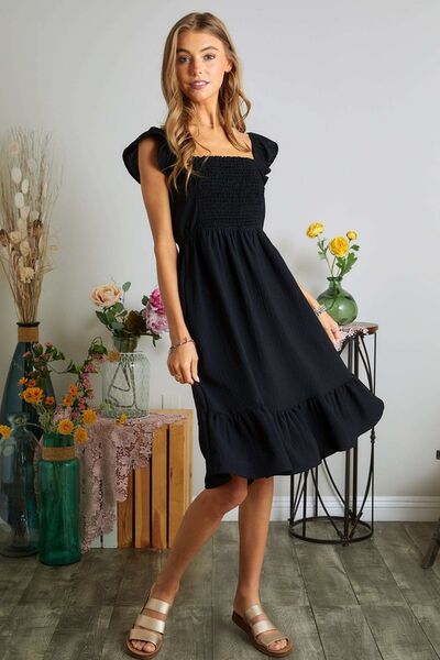 A woman wearing a black smocked square neck dress with ruffled cap sleeves, styled with subtle jewelry and surrounded by floral decor.