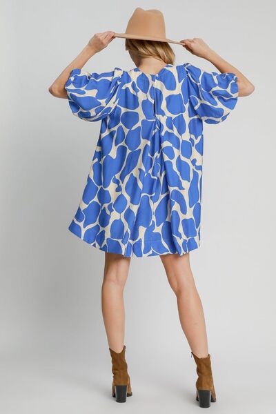 Back view of a woman wearing a blue and beige two-tone abstract print dress with puff sleeves, styled with a tan wide-brim hat for a chic and modern look.