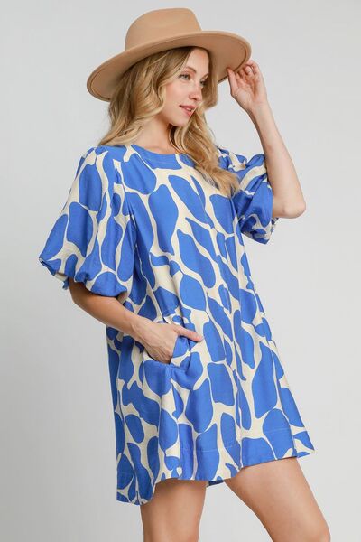 Side view of a woman wearing a blue and beige two-tone abstract print dress with puff sleeves, styled with a tan wide-brim hat for a chic and modern look.